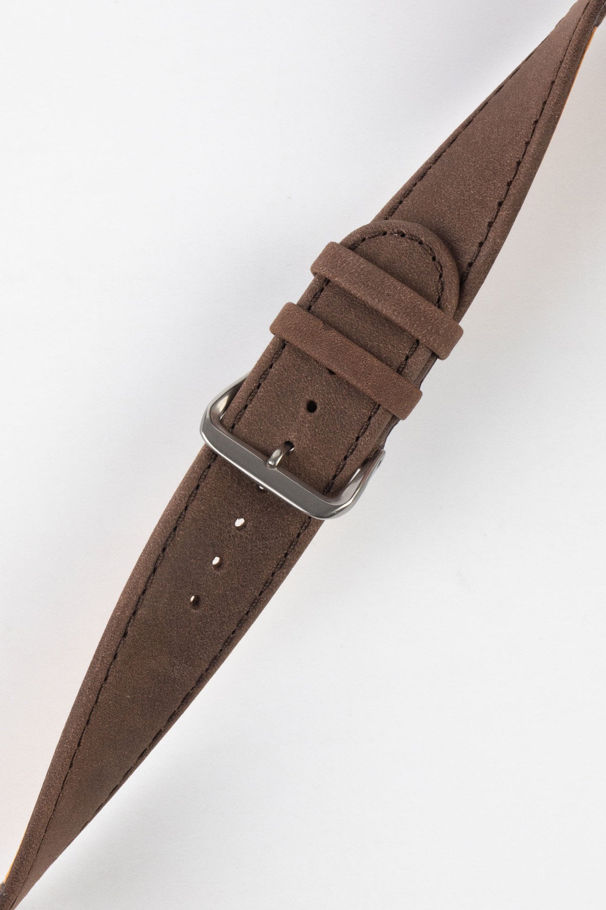 Close-up of a brown nubuck leather watch strap with a soft matte finish, tonal brown stitching, dual keeper loops, multiple adjustment holes, and a polished stainless steel buckle, displayed diagonally on a light grey background to highlight the tapered profile, vintage-inspired texture, and handcrafted leather construction.