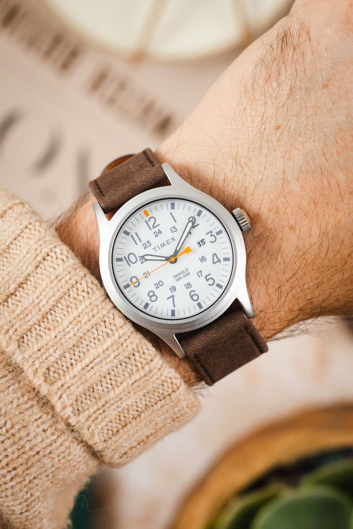 Timex Expedition field watch worn on the wrist, featuring a white dial with grey Arabic numerals, orange accent markers and seconds hand, brushed silver-tone case, and a brown nubuck leather watch strap with tonal stitching, paired with a cream knitted sweater cuff to highlight the strap's soft suede-like texture, minimalist design, and comfortable everyday style.
