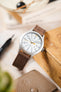 Timex Expedition field watch with a white dial, grey Arabic numerals, orange seconds hand, brushed silver-tone case, and a brown nubuck leather watch strap with tonal stitching, displayed against a vintage wooden box alongside a pen and leather accessories, highlighting the strap's soft suede-like texture, minimalist design, and casual everyday style.
