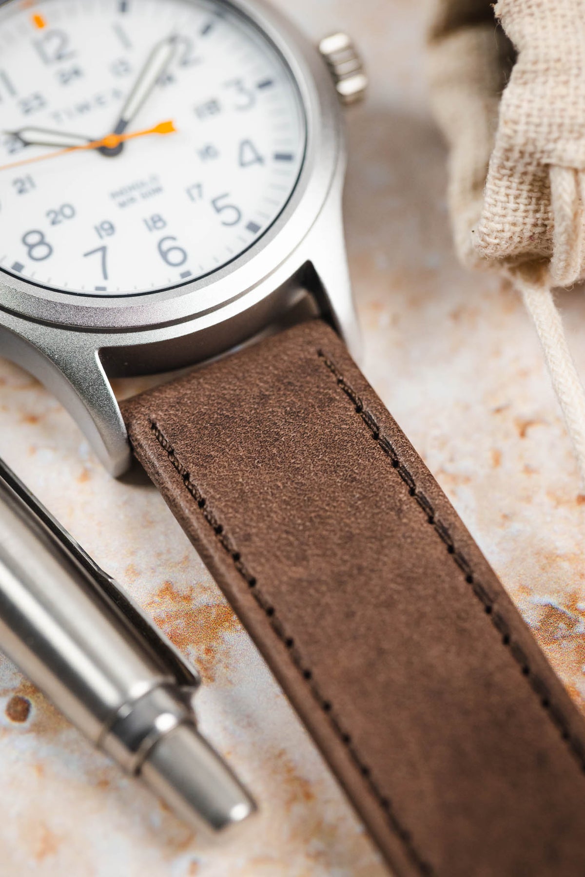 Close-up detail of a Timex Expedition field watch fitted with a brown nubuck leather watch strap, showcasing the strap's soft matte texture, tonal brown stitching, and clean minimalist construction at the lugs, displayed alongside a metal pen on a stone-effect surface to highlight the premium handcrafted leather finish and vintage-inspired character.
