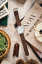 Timex Expedition field watch with a white dial, black Arabic numerals, orange accent markers, and a brown nubuck leather watch strap, displayed across a copy of NOW: The Watch Buying Guide alongside a Watch Obsession spring bar tool, succulent plant, leather accessories, and decorative pine cones, highlighting the strap's soft vintage-inspired texture, minimalist design, and versatile everyday styling.
