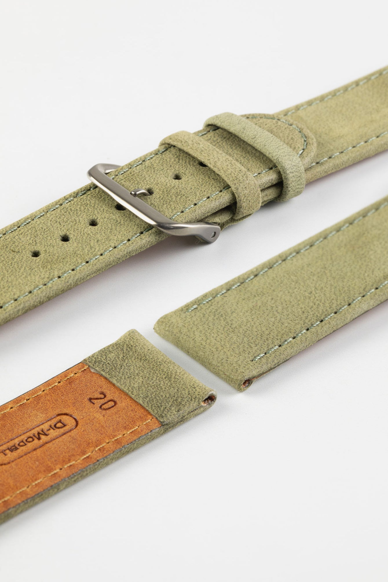 Close-up detail view of a sage green nubuck leather watch strap featuring a soft matte finish, tonal green stitching, dual keeper loops, multiple adjustment holes, a polished stainless steel buckle, and a natural tan leather lining stamped with size markings, displayed with one strap section detached on a light grey background to highlight the supple suede-like texture, contrasting underside, and premium handcrafted construction.