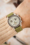 Omega x Swatch Speedmaster MoonSwatch Mission on Earth chronograph watch worn on the wrist, featuring a cream dial with brown chronograph sub-dials, brown tachymeter bezel, bioceramic case, and a sage green nubuck leather watch strap with tonal stitching, paired with a cream knitted sweater cuff to highlight the strap's soft suede-like texture, premium handcrafted construction, and complementary earth-tone colour palette.