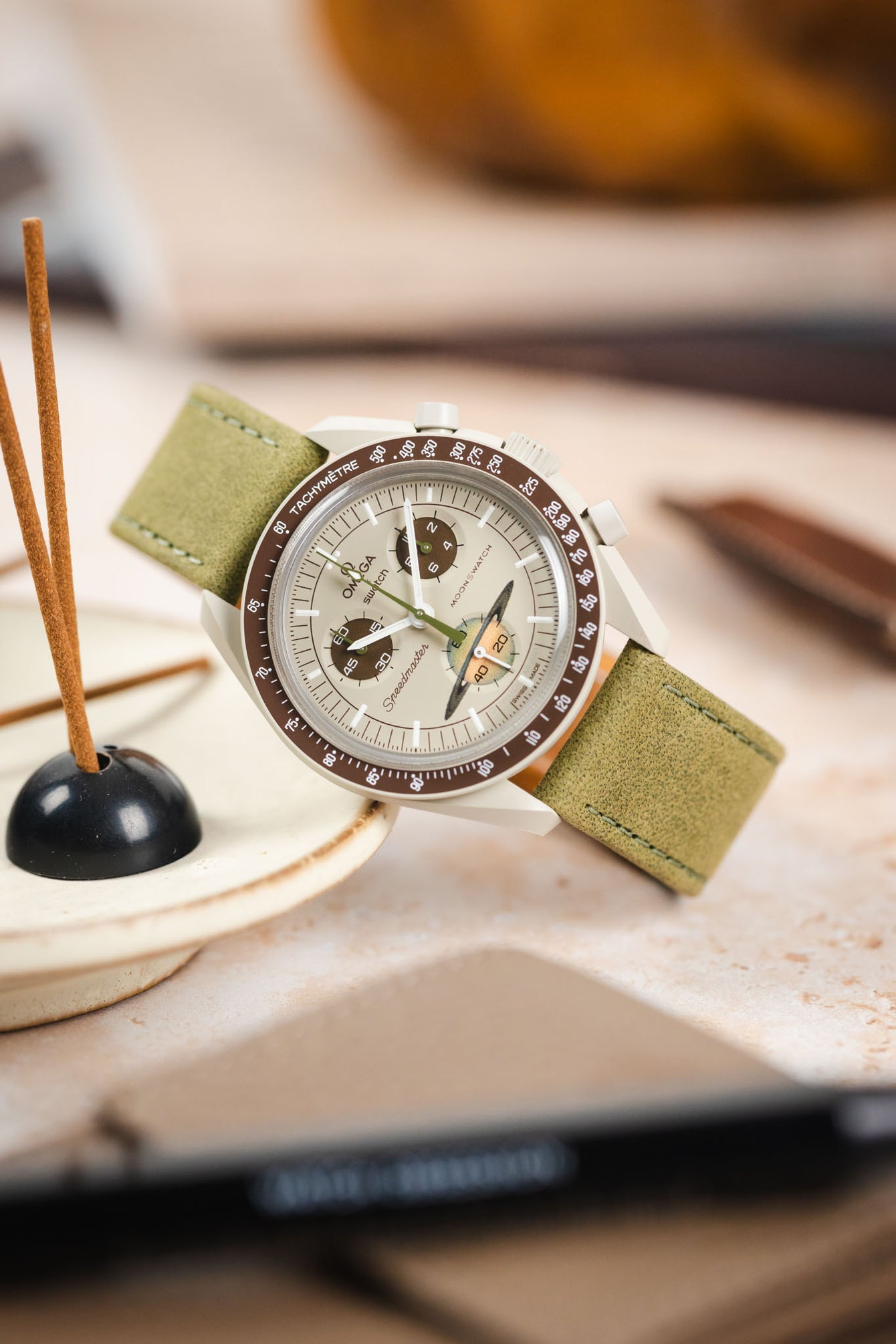 Omega x Swatch Speedmaster MoonSwatch Mission on Earth chronograph watch fitted with a sage green nubuck leather watch strap, featuring a cream dial, brown tachymeter bezel, and matching brown chronograph sub-dials, displayed beside an incense holder on a tabletop to highlight the strap's soft suede-like texture, tonal stitching, muted green colour, and premium handcrafted construction.