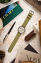 Vintage Omega Speedmaster chronograph watch fitted with a sage green nubuck leather watch strap, displayed on a stone-effect surface alongside a pen, leather accessories, books, and decorative items, highlighting the strap's soft suede-like texture, tonal stitching, muted green colour, and premium handcrafted construction.
