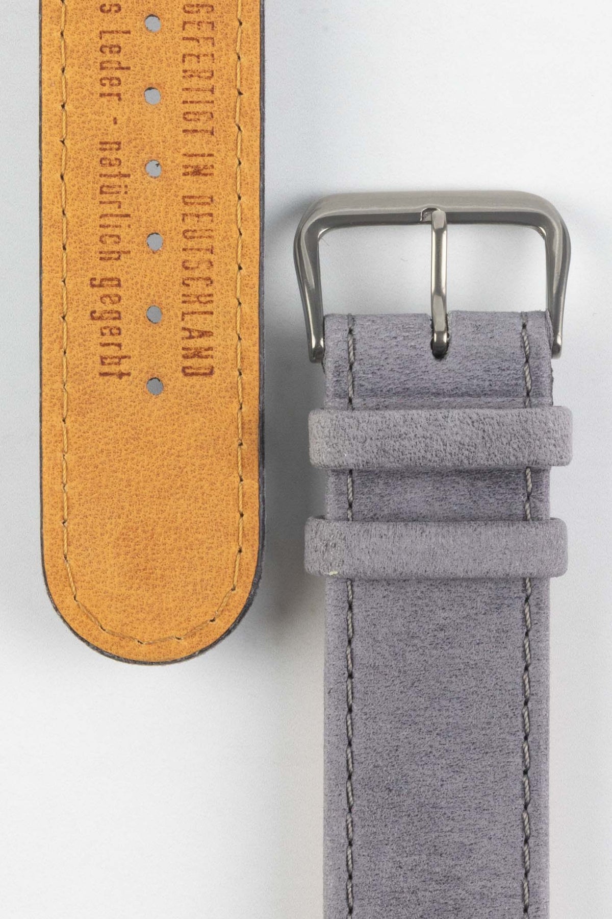 Close-up detail view of a light grey nubuck leather watch strap, showcasing a soft suede-like texture, tonal grey stitching, dual keeper loops, and a brushed stainless steel buckle, displayed alongside a natural tan leather lining embossed with “Handgefertigt in Deutschland” on a light grey background to highlight the premium handcrafted construction, contrasting underside, and vintage-inspired finish.
