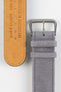 Close-up detail view of a light grey nubuck leather watch strap, showcasing a soft suede-like texture, tonal grey stitching, dual keeper loops, and a brushed stainless steel buckle, displayed alongside a natural tan leather lining embossed with “Handgefertigt in Deutschland” on a light grey background to highlight the premium handcrafted construction, contrasting underside, and vintage-inspired finish.