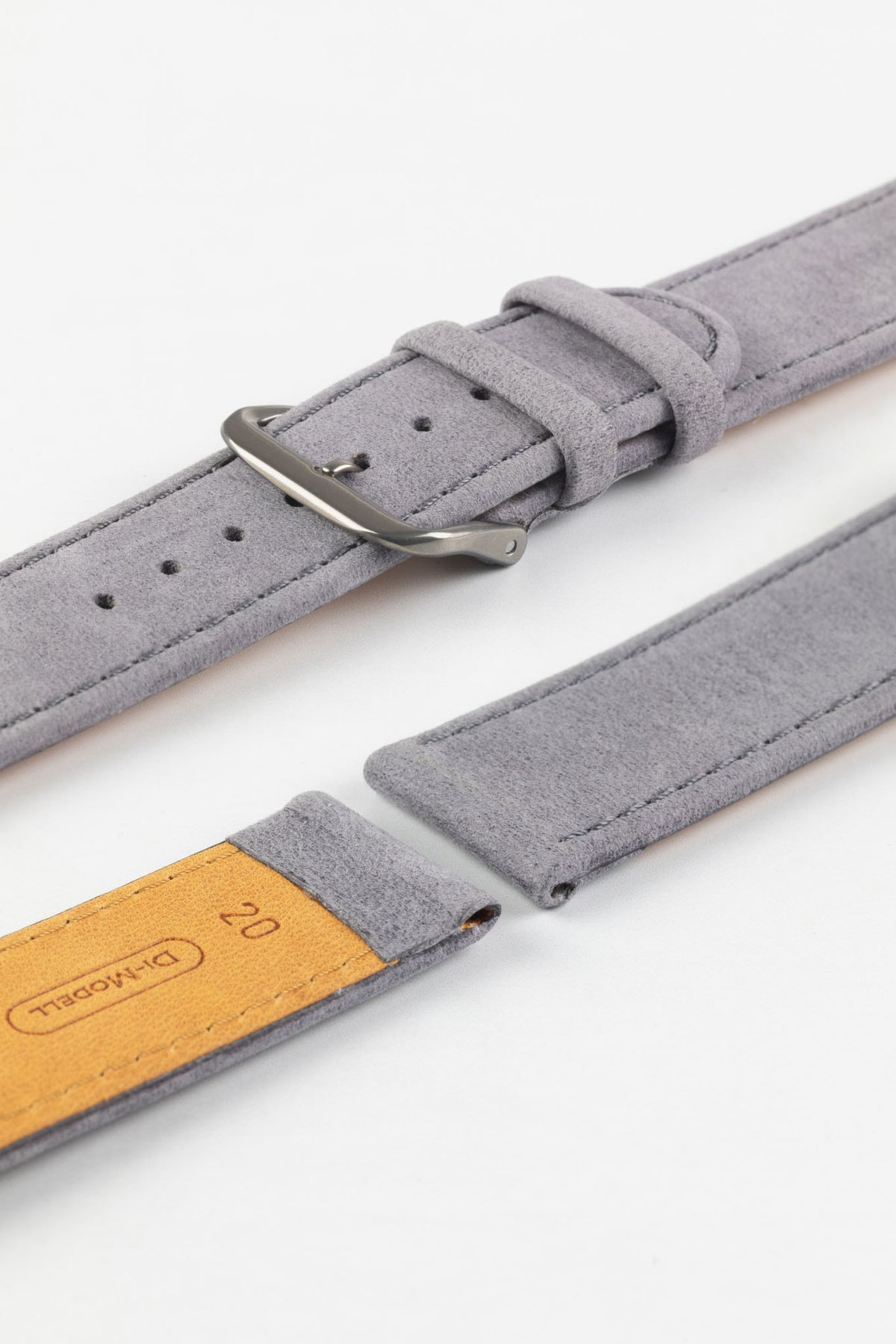Close-up detail view of a light grey nubuck leather watch strap displayed with one strap section detached, featuring a soft suede-like texture, tonal grey stitching, dual keeper loops, multiple adjustment holes, a brushed stainless steel buckle, and a natural tan leather lining embossed with the Di-Modell logo and size marking, shown on a light grey background to highlight the premium handcrafted construction and quick-release spring bar compatibility.