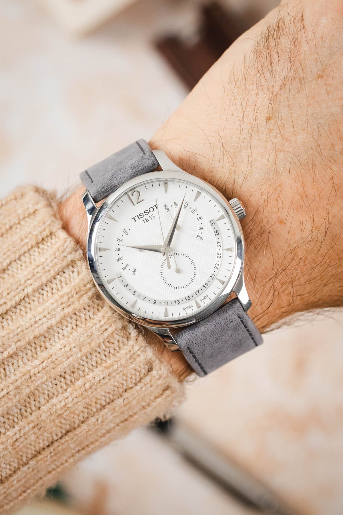 Tissot dress watch worn on the wrist, featuring a white multi-function dial, polished stainless steel case, and a light grey nubuck leather watch strap with tonal stitching and a soft suede-like texture, paired with a cream knitted sweater cuff to highlight the strap's premium handcrafted construction, minimalist styling, and refined contemporary aesthetic.