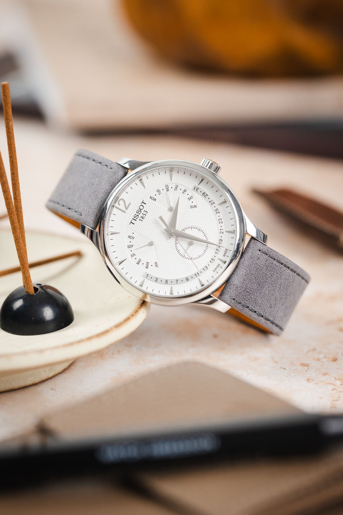 Tissot dress watch with a white multi-function dial and polished stainless steel case, fitted with a light grey nubuck leather watch strap featuring tonal stitching and a soft suede-like texture, displayed beside an incense holder on a stone-effect surface to highlight the strap's premium handcrafted construction, minimalist design, and refined contemporary styling.
