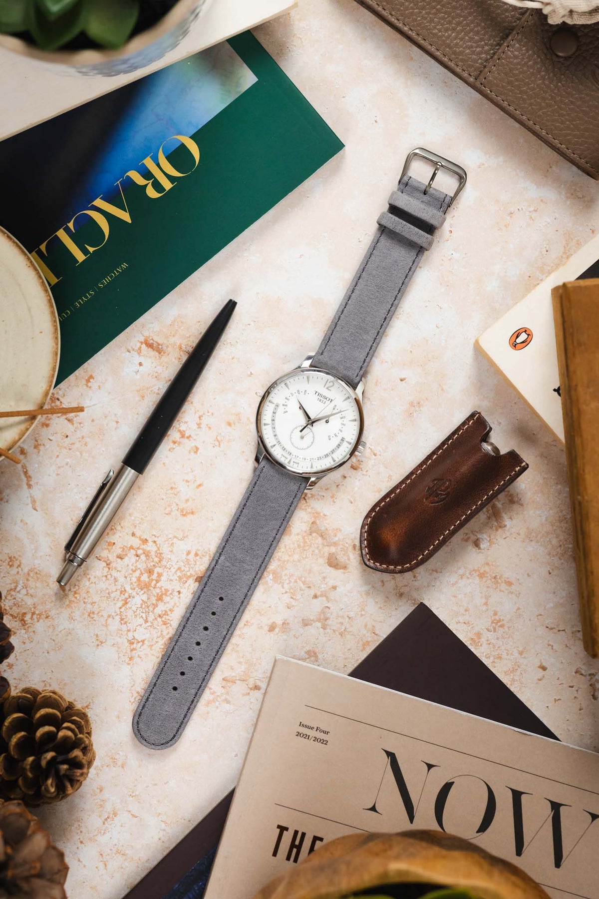 Tissot dress watch with a white dial and polished stainless steel case, fitted with a light grey nubuck leather watch strap, displayed on a stone-effect surface alongside an Oracle Time magazine, pen, leather accessories, books, and decorative items, highlighting the strap's soft suede-like texture, tonal stitching, minimalist design, and premium handcrafted construction.