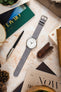Tissot dress watch with a white dial and polished stainless steel case, fitted with a light grey nubuck leather watch strap, displayed on a stone-effect surface alongside an Oracle Time magazine, pen, leather accessories, books, and decorative items, highlighting the strap's soft suede-like texture, tonal stitching, minimalist design, and premium handcrafted construction.