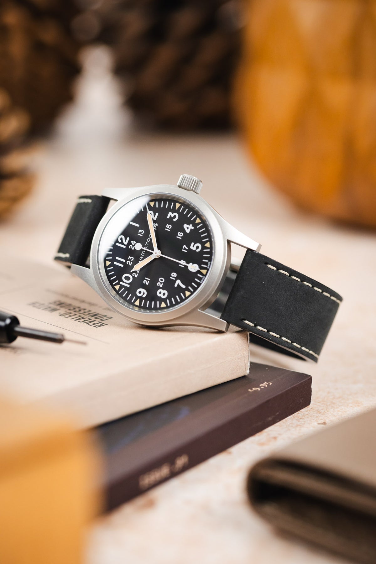 Hamilton Khaki Field watch with a black dial, white Arabic numerals, luminous syringe-style hands, and a brushed stainless steel case, fitted with a black leather watch strap featuring contrasting cream stitching. Displayed at an angle on stacked books beside a Watch Obsession spring bar tool, the image highlights the strap's smooth matte finish, vintage-inspired design, and premium handcrafted construction.