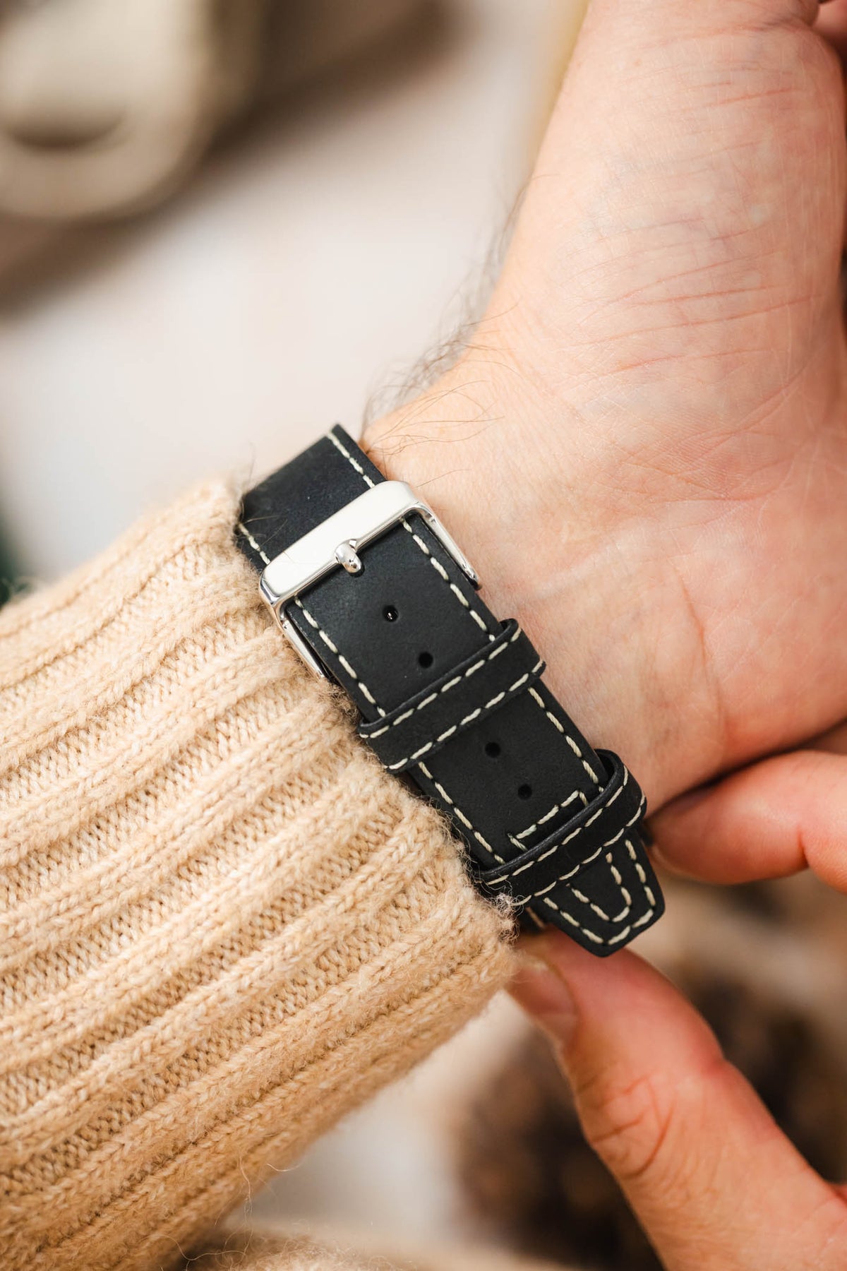 Close-up of a black leather watch strap worn on the wrist, featuring contrasting cream stitching, dual keeper loops, multiple adjustment holes, and a polished stainless steel buckle, shown with a cream knitted sweater cuff to highlight the strap's smooth matte finish, premium handcrafted construction, secure fit, and vintage-inspired design.