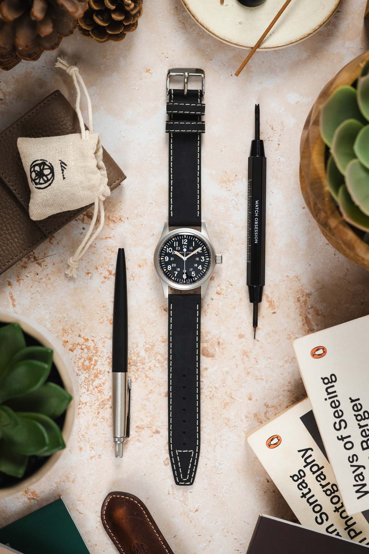 Hamilton Khaki Field watch with a black dial, white Arabic numerals, and brushed stainless steel case, fitted with a black leather watch strap featuring contrasting cream stitching and a vintage-inspired tapered design. Displayed on a stone-effect surface alongside a Watch Obsession spring bar tool, pen, leather accessories, books, succulents, and decorative pine cones, highlighting the strap’s premium handcrafted construction and classic field-watch styling.