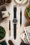 Hamilton Khaki Field watch with a black dial, white Arabic numerals, and brushed stainless steel case, fitted with a black leather watch strap featuring contrasting cream stitching and a vintage-inspired tapered design. Displayed on a stone-effect surface alongside a Watch Obsession spring bar tool, pen, leather accessories, books, succulents, and decorative pine cones, highlighting the strap’s premium handcrafted construction and classic field-watch styling.