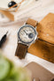 Studio Underd0g Bicompax mechanical chronograph watch with a textured cream dial, contrasting dark grey and off-white sub-dials, polished stainless steel case, and a dark brown leather watch strap featuring cream contrast stitching and a vintage-inspired finish, displayed against a wooden box alongside a spring bar tool and leather accessories, highlighting the strap's premium handcrafted construction and classic heritage styling.
