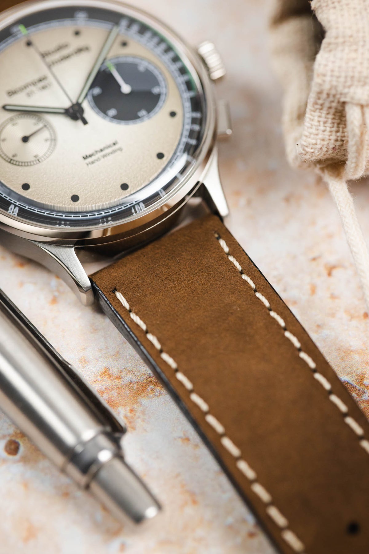 Close-up detail of a vintage-style chronograph watch fitted with a dark brown leather watch strap featuring cream contrast stitching and a smooth matte finish, displayed beside a metal pen on a stone-effect surface. The image highlights the strap's supple leather texture, tapered profile, premium handcrafted construction, and classic heritage-inspired styling.