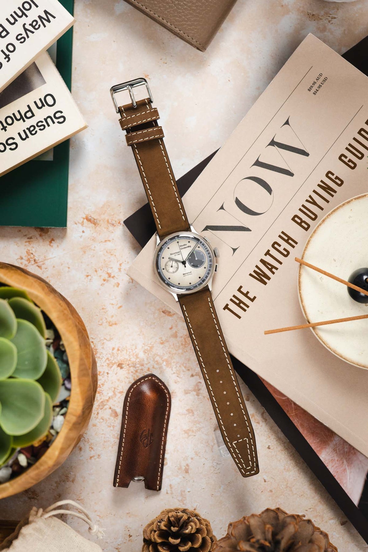 Vintage-style Studio Underd0g Bicompax chronograph watch with a cream dial, contrasting dark blue and cream sub-dials, and a dark brown leather watch strap featuring cream contrast stitching and a tapered pointed tip, displayed across a copy of NOW: The Watch Buying Guide alongside a leather spring bar tool sleeve, succulent plant, incense holder, books, and decorative pine cones, highlighting the strap's premium handcrafted construction and classic vintage-inspired styling.
