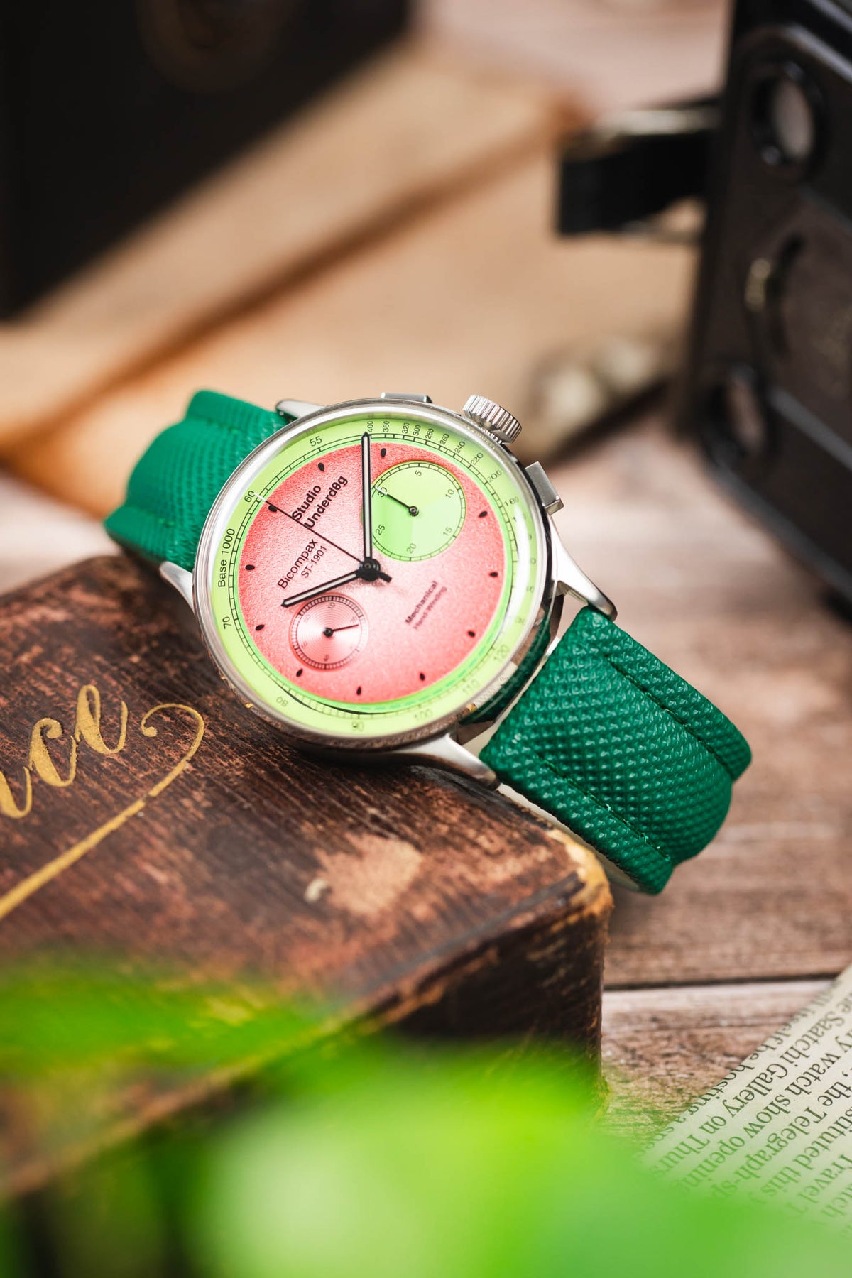 Studio Underd0g chronograph watch on a green textured leather strap, displayed on a vintage wooden box with retro camera accessories.