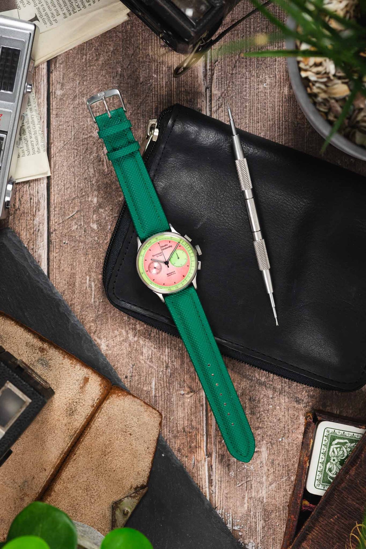 Studio Underd0g chronograph watch on a green textured leather strap, displayed with vintage accessories and watchmaking tools on a rustic wooden surface.