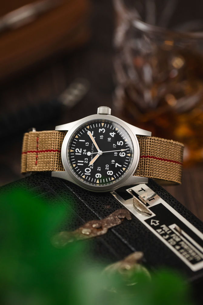 Black dial field watch leant on a vintage camera fitted to a brown parachute webbing watch strap with brushed stainless steel hardware
