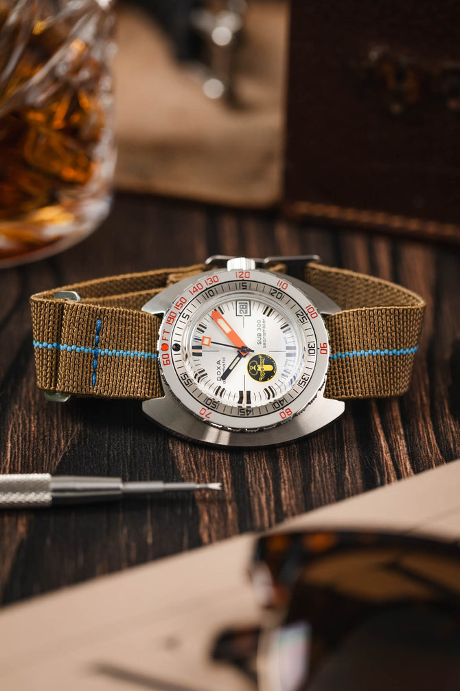 Dive watch on a wooden table fitted to a brown parachute webbing watch strap with brushed stainless steel hardware