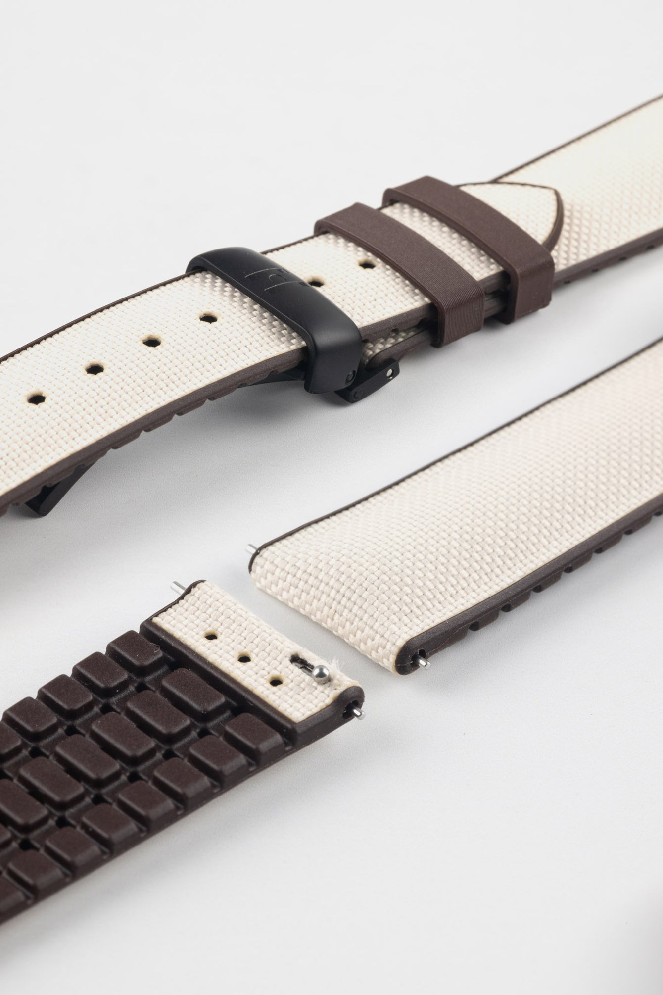 Beige textile-effect watch strap with brown rubber backing and a black deployment clasp, shown in separate sections on a white background. Features a woven-look surface, brown edges, and a patterned rubber underside.