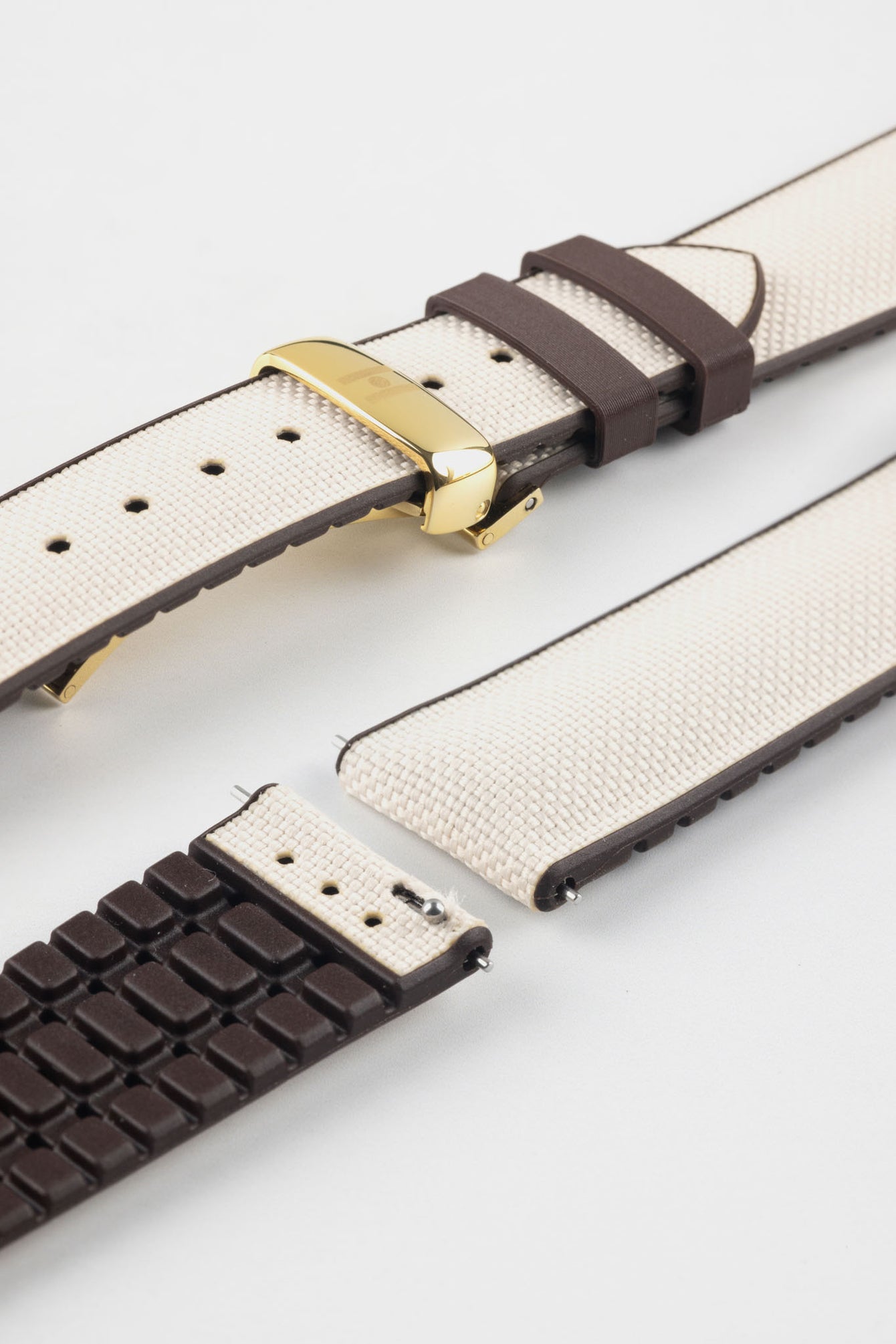 Beige textile-effect watch strap with brown rubber backing and a gold-tone deployment clasp, shown in separate sections on a white background. Features a woven-look surface, brown edges, and a patterned rubber underside.