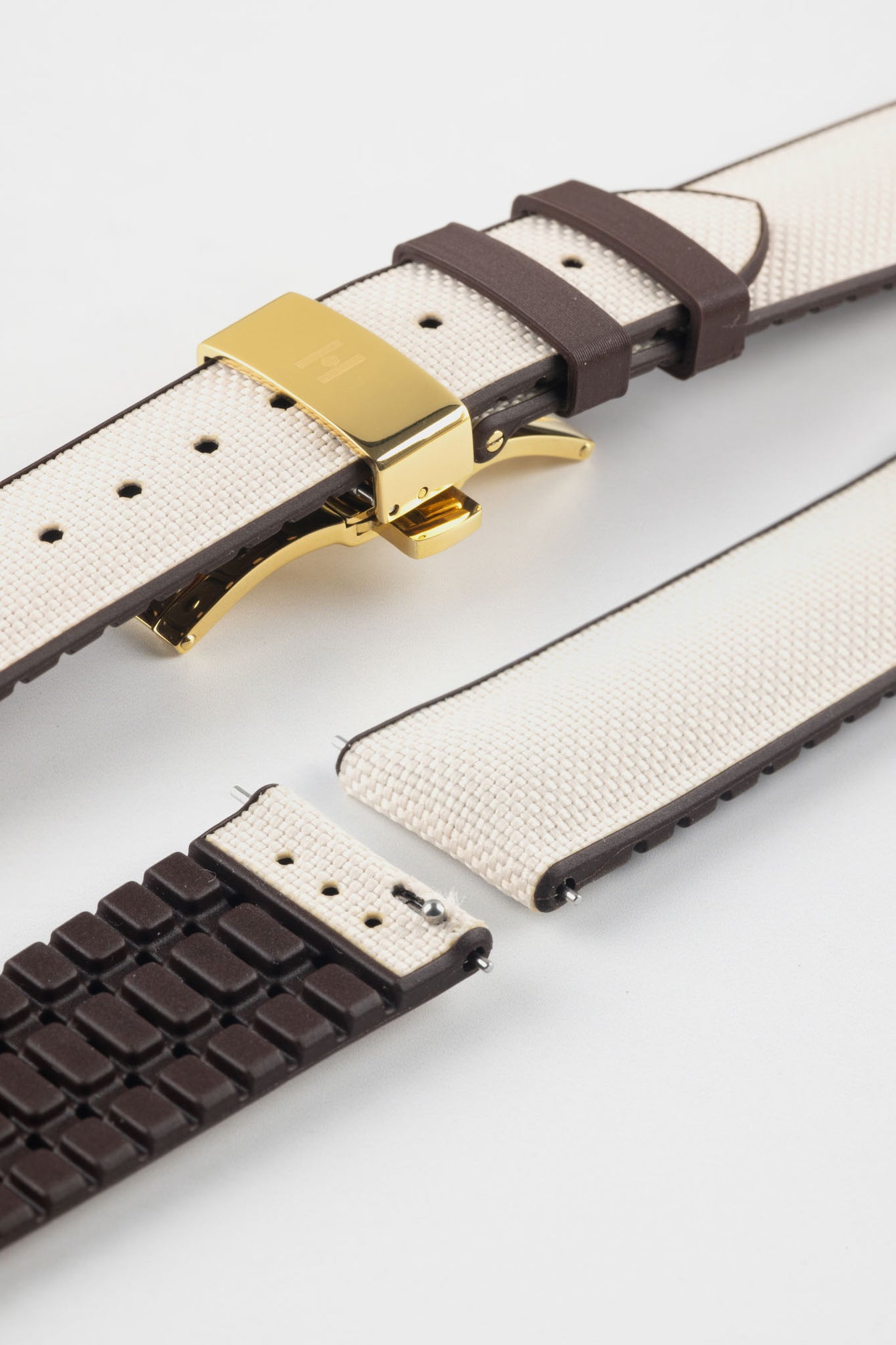 Beige textile-effect watch strap with brown rubber backing and a gold-tone deployment clasp, shown in separate sections on a white background. Features a woven-look surface, brown edges, and a patterned rubber underside.