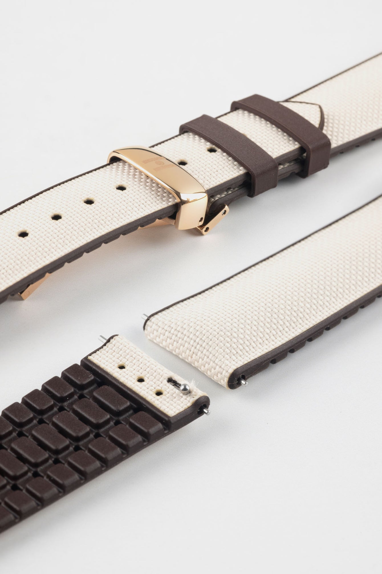 Beige textile-effect watch strap with brown rubber backing and a rose gold-tone deployment clasp, shown in separate sections on a white background. Features a woven-look surface, brown edges, and a patterned rubber underside.