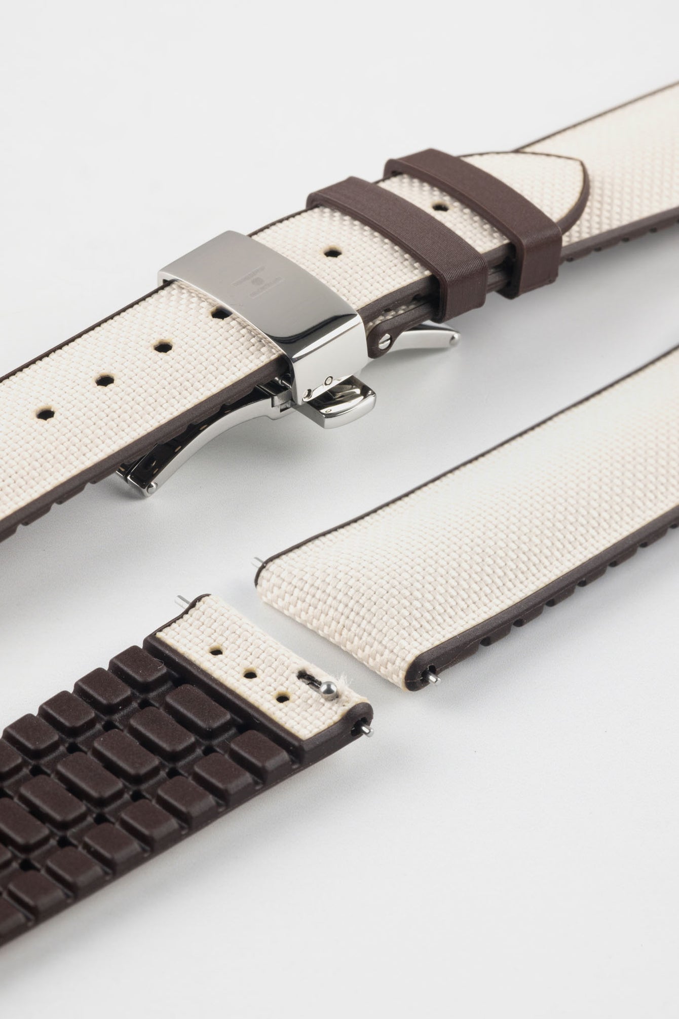Beige textile-effect watch strap with brown rubber backing and a silver-tone deployment clasp, shown in separate sections on a white background. Features a woven-look surface, brown edges, and a patterned rubber underside.