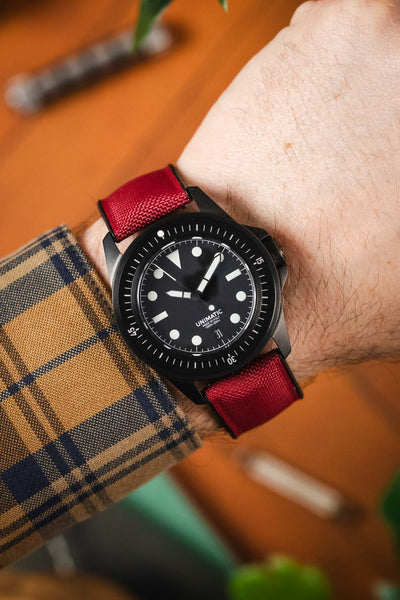 A close-up shot of a person's wrist wearing a black Unimatic Modello Tre watch with a vibrant red Hirsch Arne Performance strap. The watch features a black dial with white markers and hands, and the strap has a subtle textured pattern. The person is wearing a blue and gold plaid shirt, and a blurred wooden surface is visible in the background.