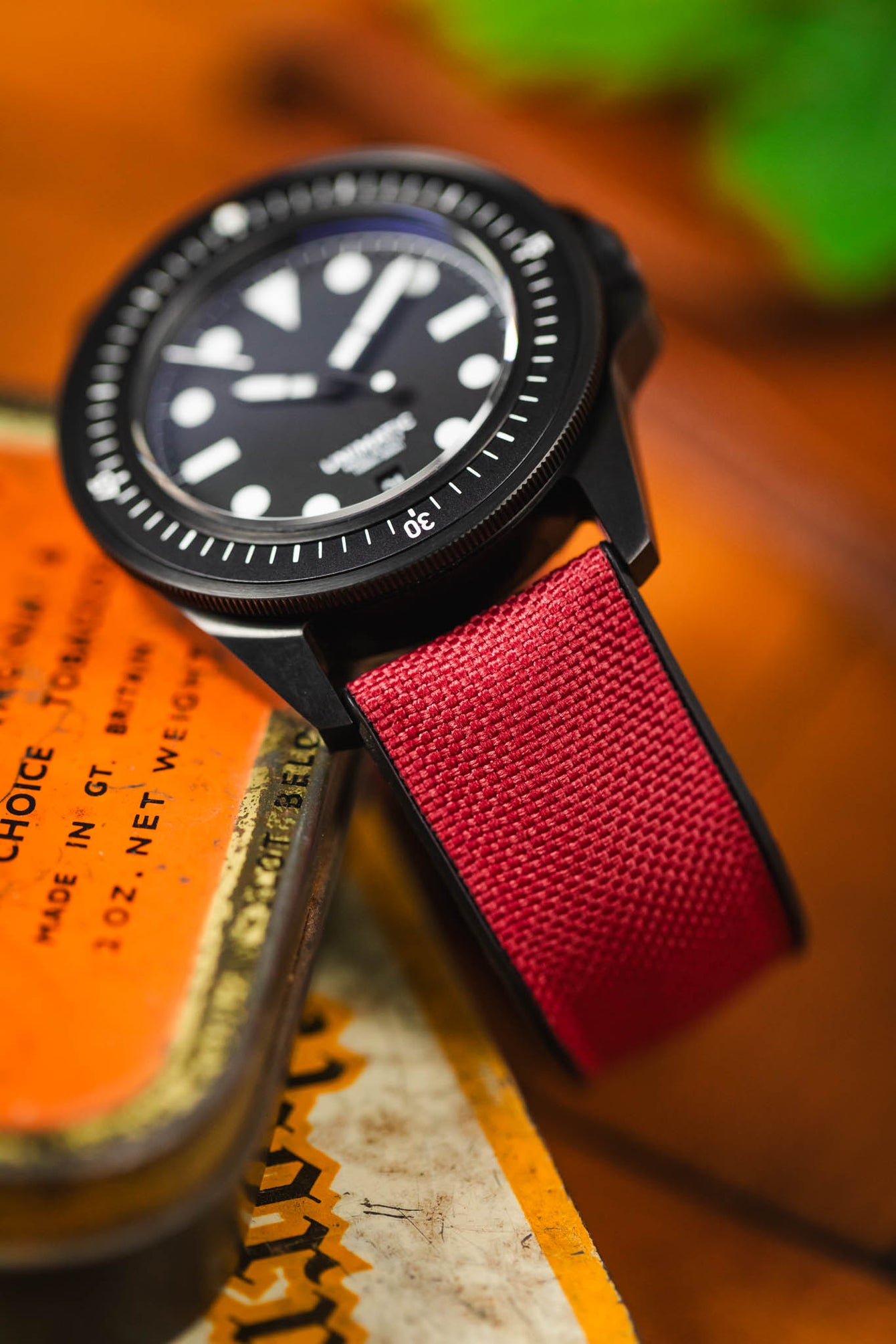 A close-up shot of a black Unimatic watch with a distinctive burgundy Hirsch Arne sailcloth performance strap. The watch face is partially visible, resting on a vintage orange and gold tin. The textured weave of the red strap is clearly in focus.