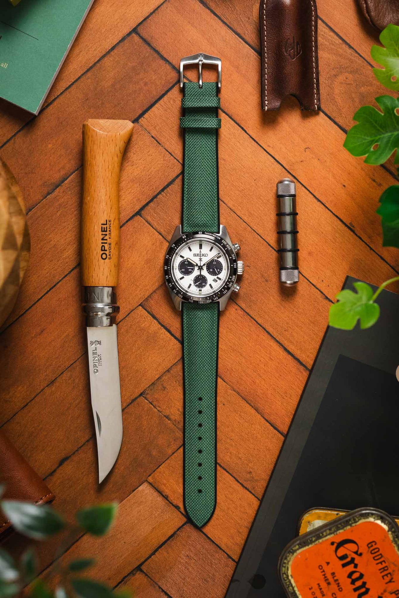 A top-down view of a silver Seiko Speedtimer chronograph watch with a dark green Hirsch Arne Animal-Free Sailcloth Effect Performance strap, laid out on a wooden herringbone surface amongst various EDC (Everyday Carry) items. The watch has a white dial with black subdials and a silver pin buckle. Other items include a closed Opinel folding knife with a wooden handle, a brown leather pouch, a metal accessory, and a glimpse of a green leafy plant and a vintage tin in the corners.