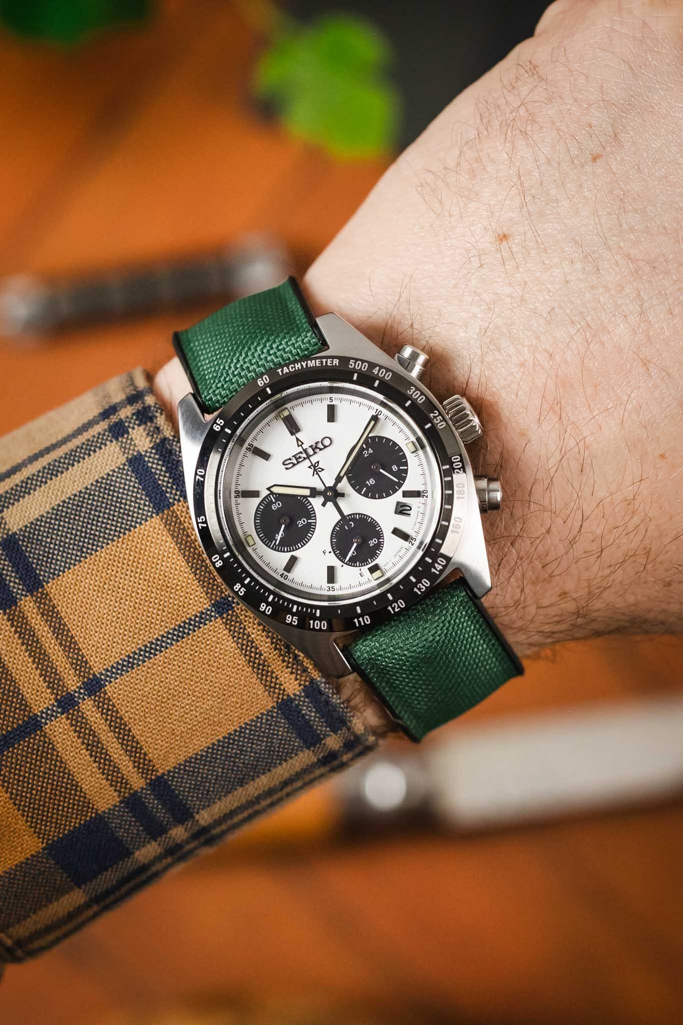 A close-up shot of a person's wrist wearing a silver Seiko Speedtimer chronograph watch with a dark green Hirsch Arne Animal-Free Sailcloth Effect Performance strap. The watch has a white dial with black subdials and a tachymeter bezel. The person is wearing a blue and gold plaid shirt, and a blurred wooden surface is visible in the background.