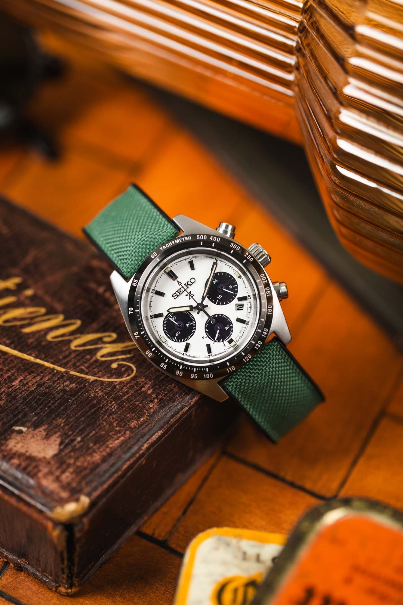A silver Seiko Speedtimer chronograph watch with a dark green Hirsch Arne Animal-Free Sailcloth Effect Performance strap is displayed on a textured brown surface, possibly a wooden box or table. The watch features a white dial with black subdials and a tachymeter bezel. The dark green textured strap is clearly visible. The background is slightly blurred, suggesting a warm, indoor setting with other objects partially visible.