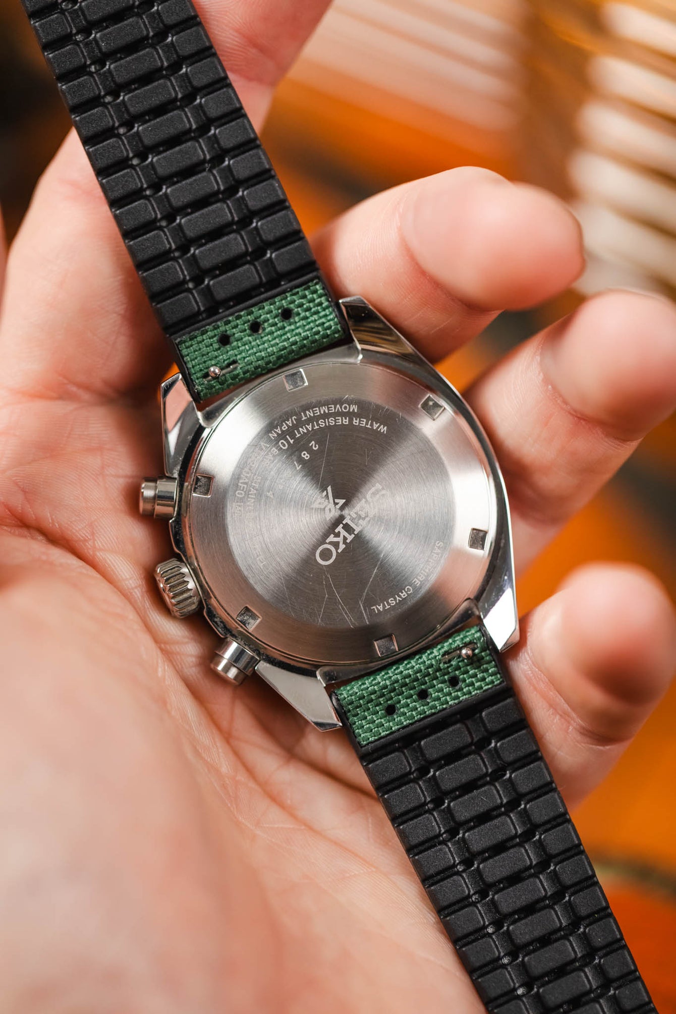 A close-up shot of the back of a silver Seiko Speedtimer chronograph watch being held in a person's hand. The watch has a visible stainless steel caseback with "SEIKO" and other technical details engraved on it. The watch is fitted with a dark green Hirsch Arne Animal-Free Sailcloth Effect Performance strap, showing the textured green fabric where it attaches to the watch case and the black rubberized underside of the strap. The person's fingers are gently holding the watch, and a blurred orange and brown b