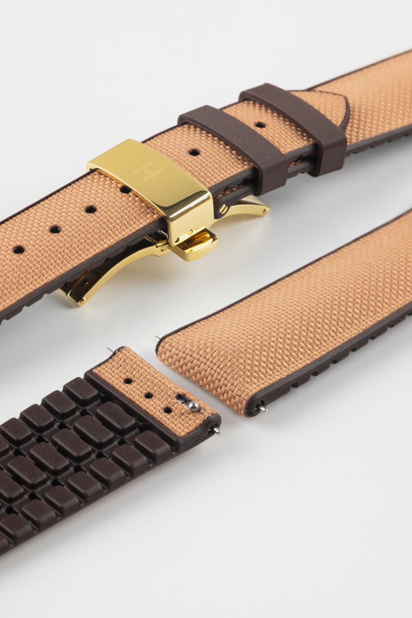 Golden brown textile-effect watch strap with brown rubber backing and a gold-tone deployment clasp, shown in separate sections on a white background. Features a woven-look surface, brown edges, and a patterned rubber underside.