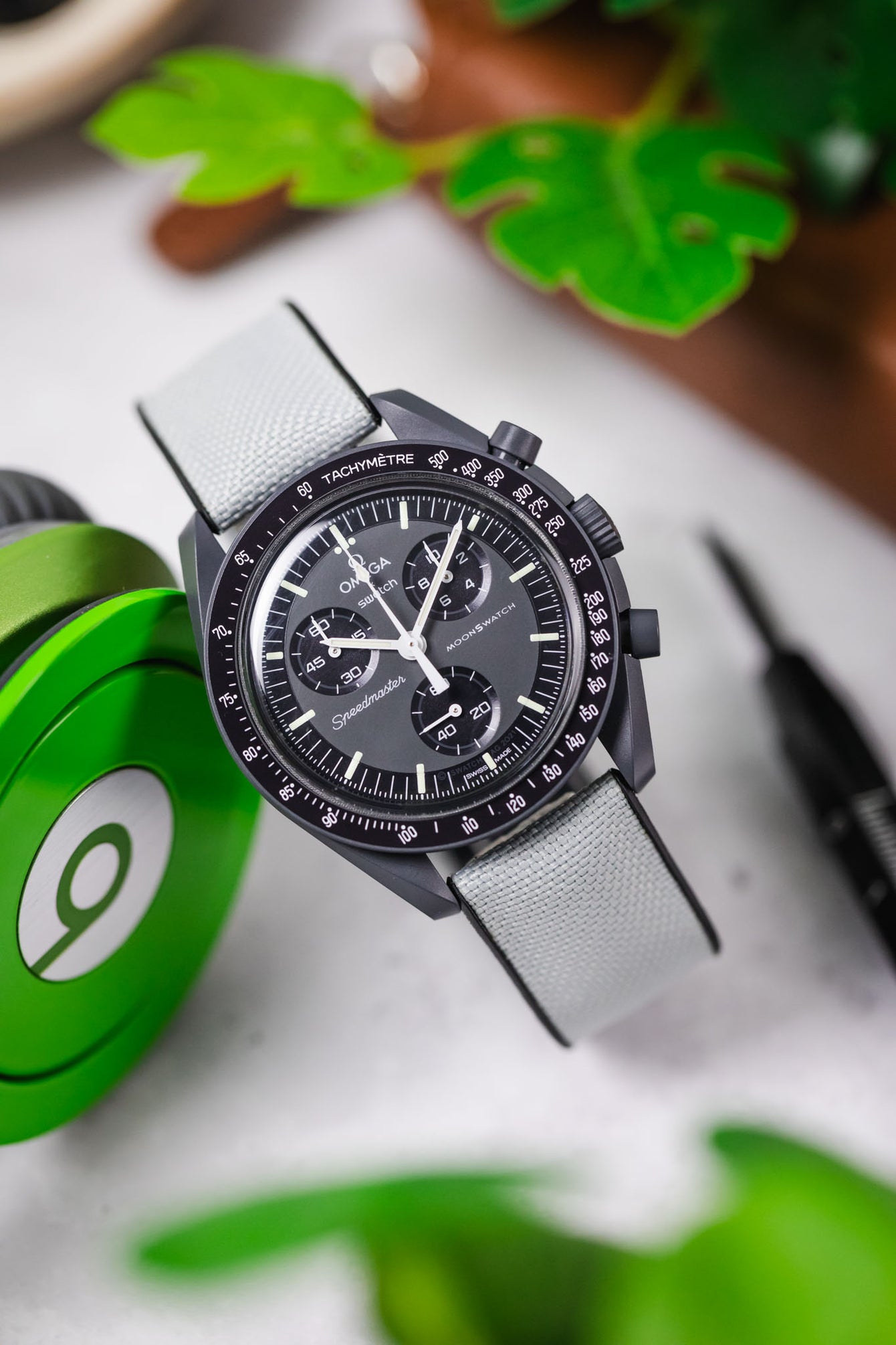  An overhead shot of a black ceramic Omega Speedmaster Dark Side of the Moon watch with a grey dial and a light grey Hirsch Arne Performance Watch Strap, laid on a white textured surface next to bright green over-ear headphones and blurred greenery.
