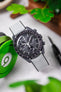  An overhead shot of a black ceramic Omega Speedmaster Dark Side of the Moon watch with a grey dial and a light grey Hirsch Arne Performance Watch Strap, laid on a white textured surface next to bright green over-ear headphones and blurred greenery.
