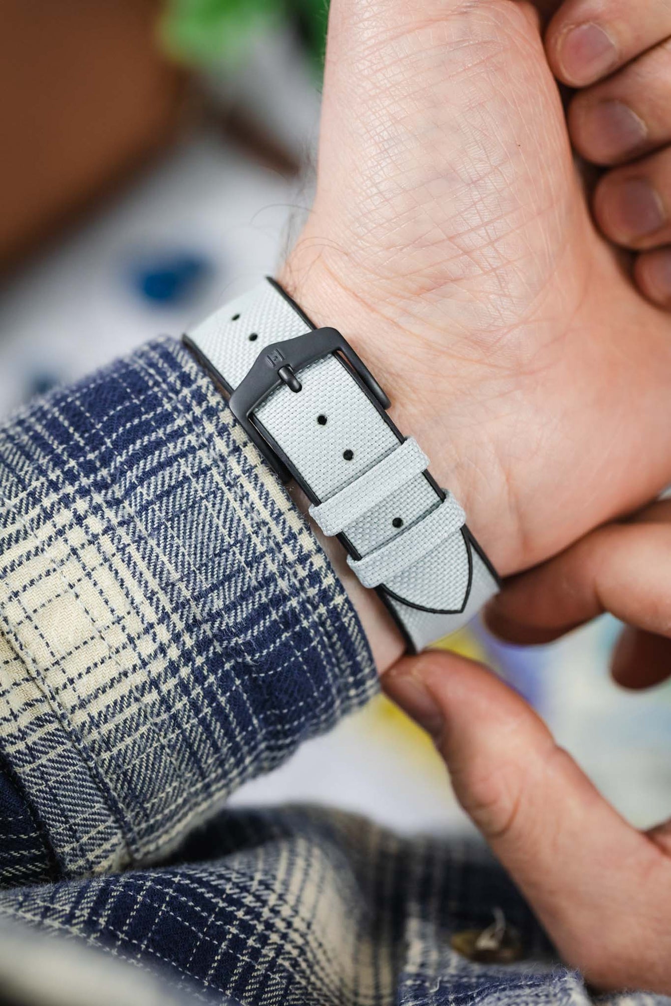 An overhead shot of a hand securing a light grey Hirsch Arne Performance Watch Strap with a black PVD tang buckle onto a wrist. The textured surface of the grey strap is visible, along with a plaid shirt cuff.