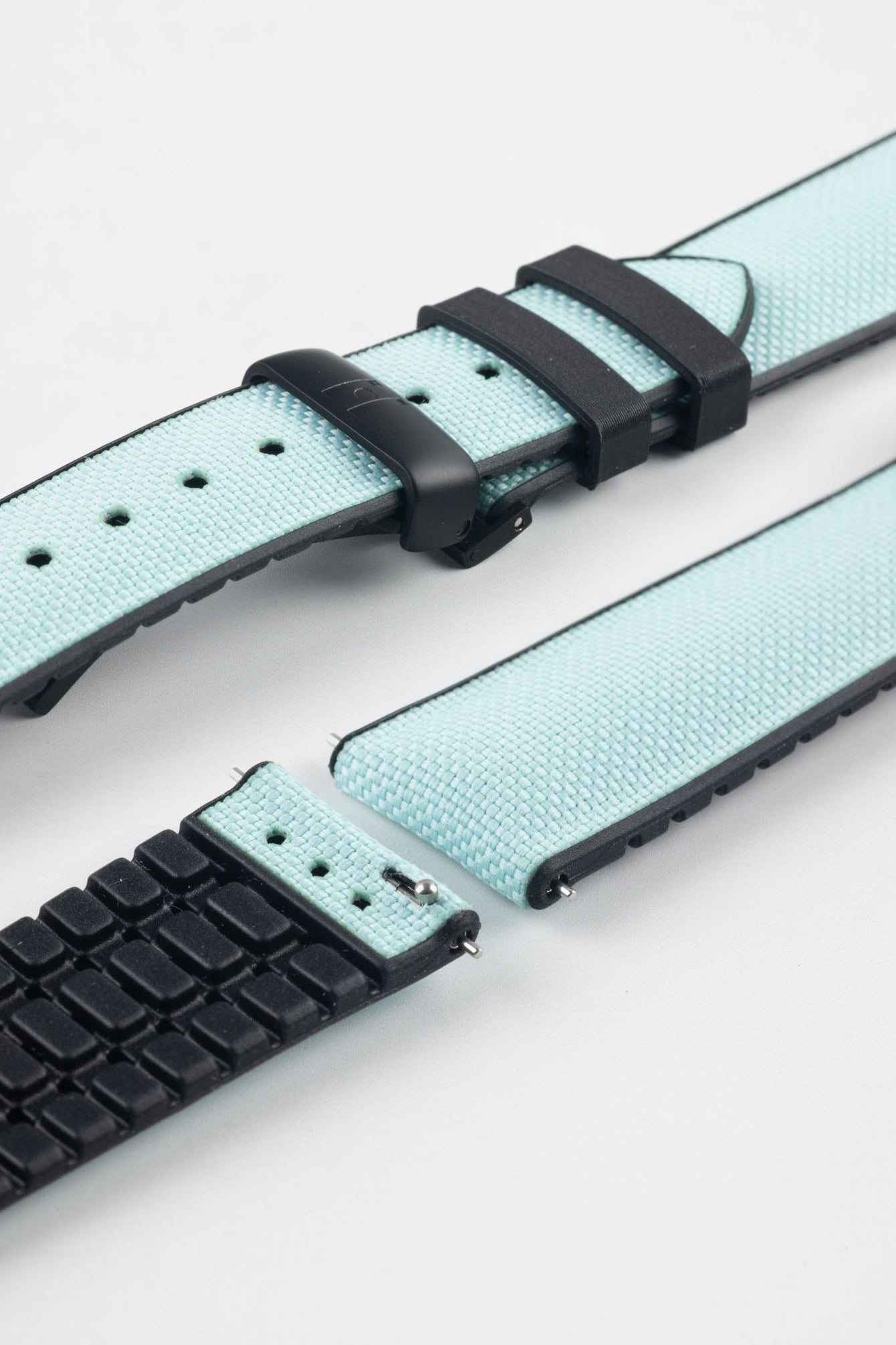 Ice blue textile-effect watch strap with black rubber backing and a black deployment clasp, shown in separate sections on a white background. Features a woven-look surface, black edges, and a patterned rubber underside.