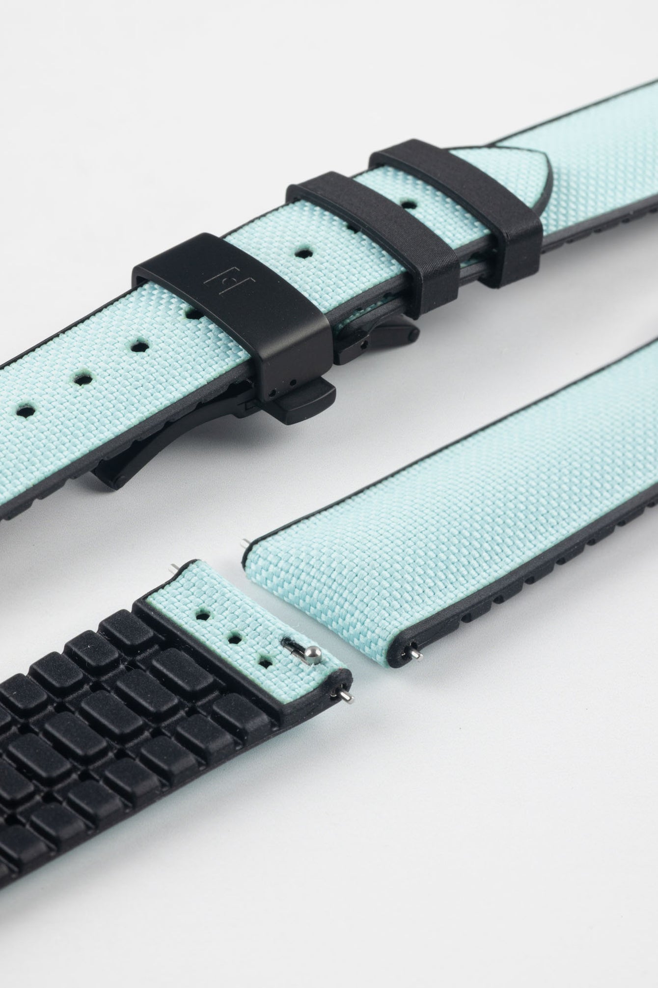 Ice blue textile-effect watch strap with black rubber backing and a black deployment clasp, shown in separate sections on a white background. Features a woven-look surface, black edges, and a patterned rubber underside.