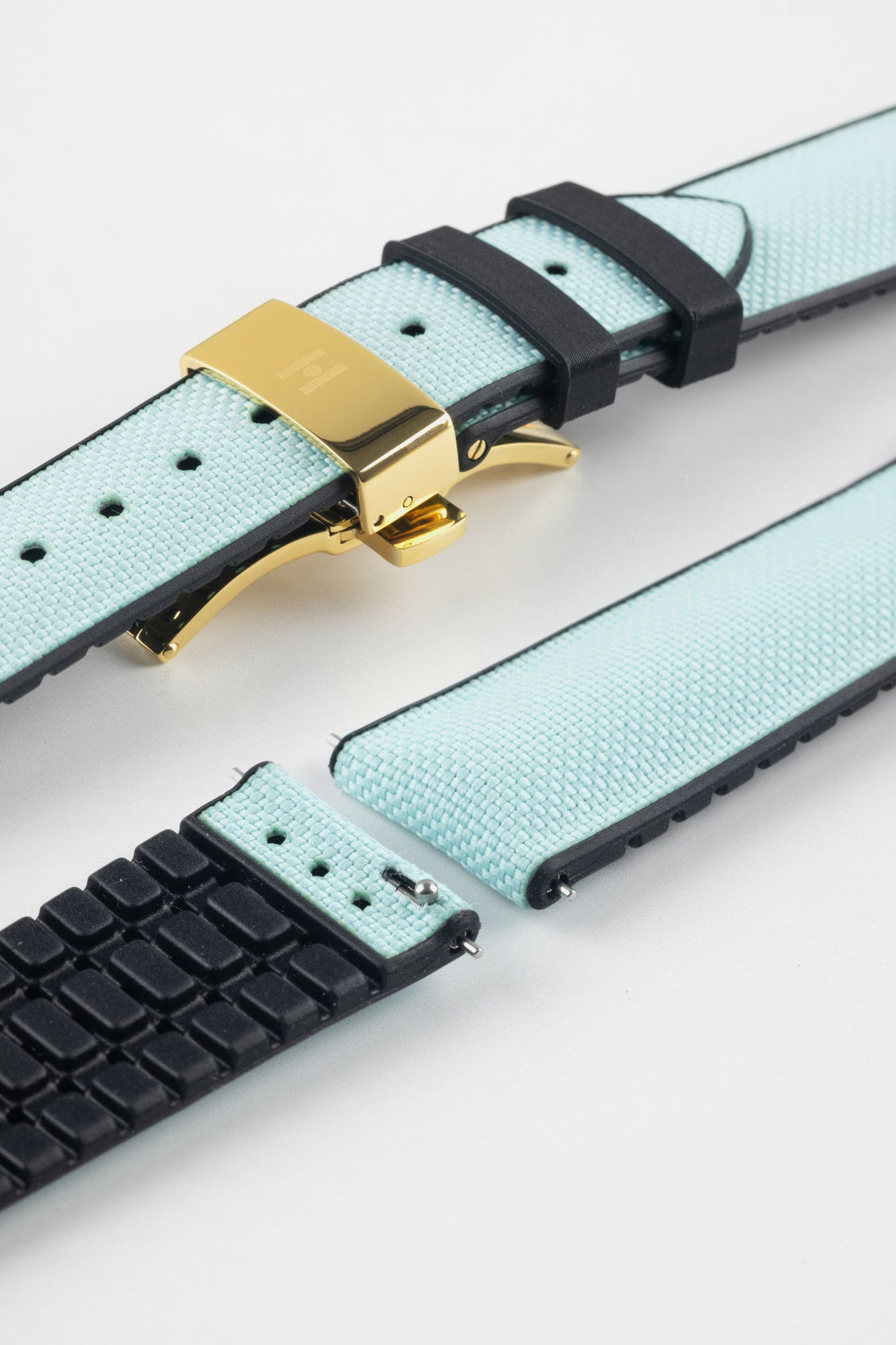 Ice blue textile-effect watch strap with black rubber backing and a gold-tone deployment clasp, shown in separate sections on a white background. Features a woven-look surface, black edges, and a patterned rubber underside.