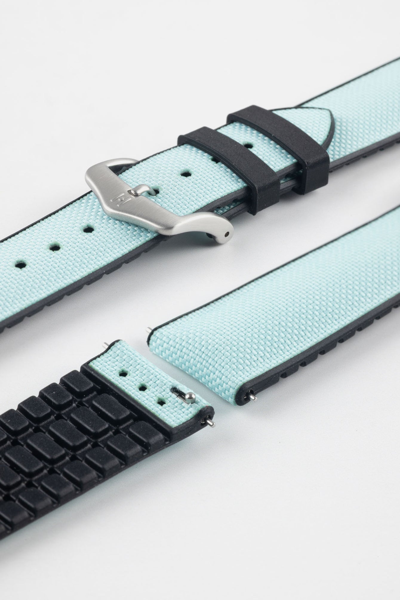 Ice blue textile-effect watch strap with black rubber backing and a brushed silver-tone buckle, shown in separate sections on a white background. Features a woven-look surface, black edges, and a patterned rubber underside.