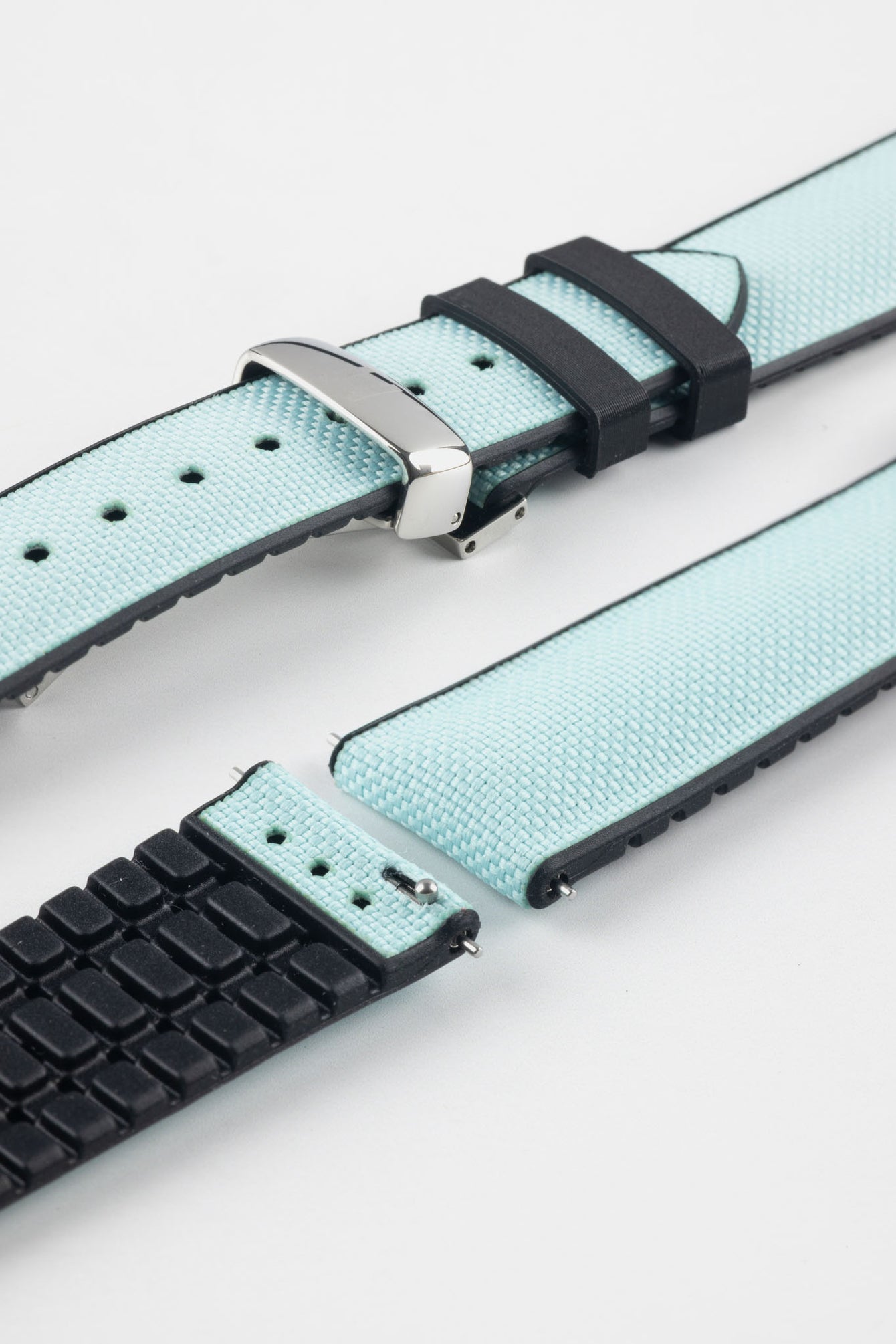 Ice blue textile-effect watch strap with black rubber backing and a silver-tone deployment clasp, shown in separate sections on a white background. Features a woven-look surface, black edges, and a patterned rubber underside.