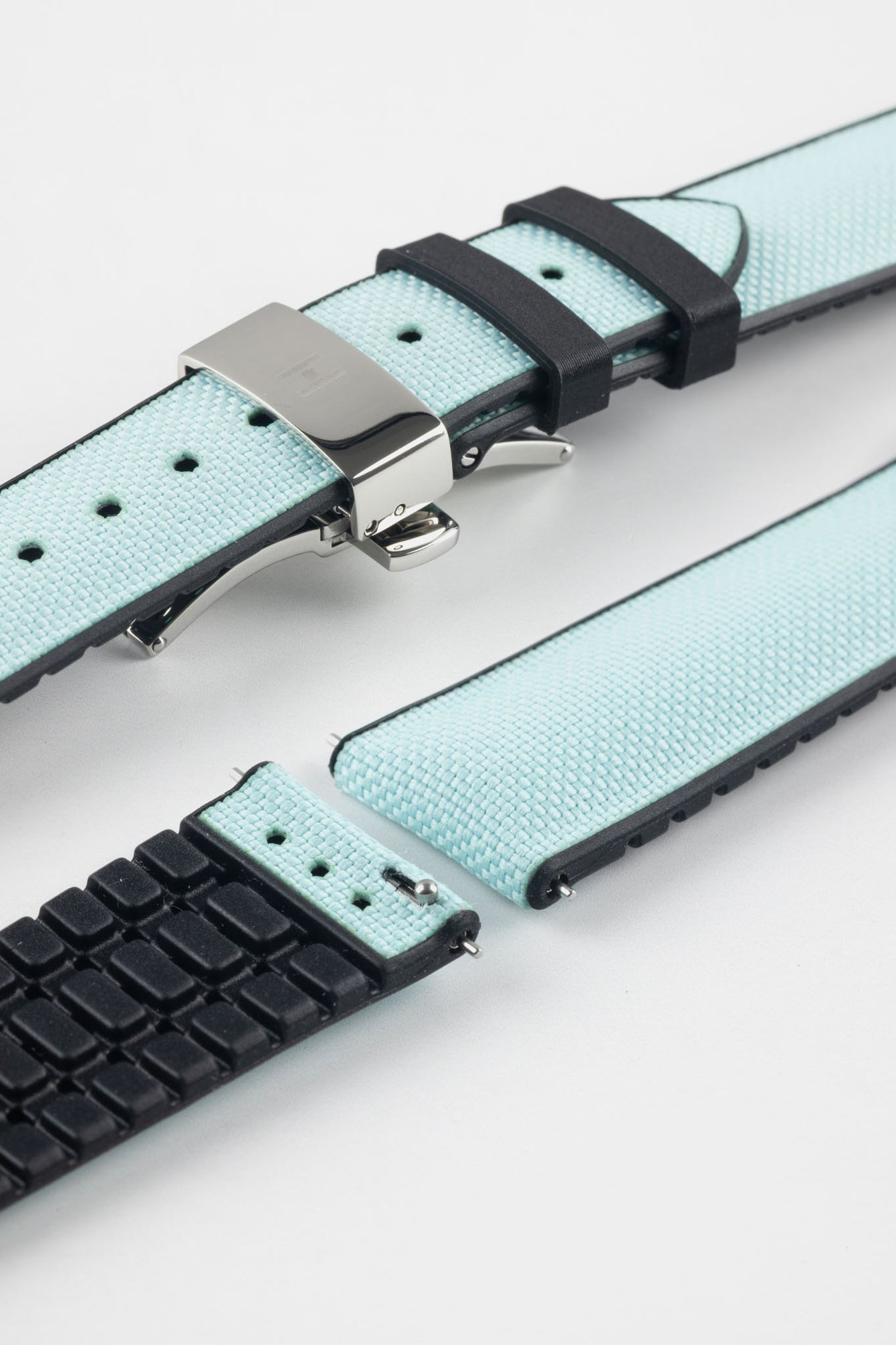 Ice blue textile-effect watch strap with black rubber backing and a silver-tone deployment clasp, shown in separate sections on a white background. Features a woven-look surface, black edges, and a patterned rubber underside.