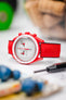 A close-up, eye-level shot of a red Omega MoonSwatch Mission to Mars watch with a white dial and a red tachymeter bezel, fitted with a red Hirsch Arne Performance Watch Strap. The watch is positioned on a white textured surface, with a black watch tool in the foreground and blurred green objects in the background.