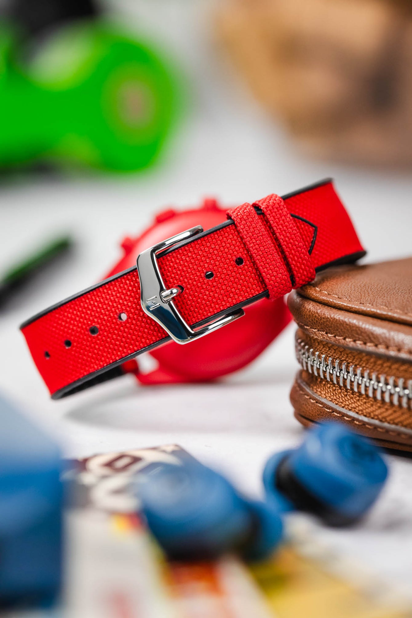 A close-up, eye-level shot of a red Omega MoonSwatch watch with a red case and white dial, fitted with a red Hirsch Arne Performance Watch Strap and a polished silver-toned tang buckle. It rests near a brown leather zippered watch case, with various blurred objects in the foreground and background.